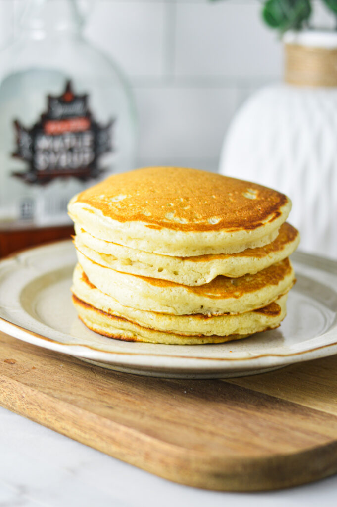 A stack of golden brown Plain Pancakes on a white plate, with a jar of maple syrup in the background.