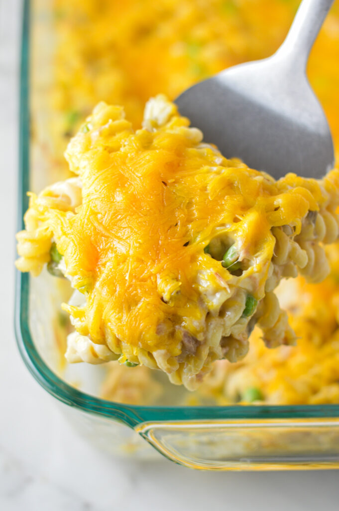 A spatula taking a scoop of Tuna Casserole from a large pyrex baking dish, showing the creamy mixture of noodles, cheese and peas.