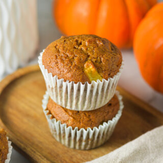 A stack of two golden brown Apple Pumpkin Muffins served on a rustic wooden tray with small pumpkins in the background.