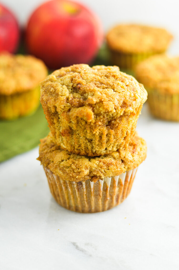 Two Apple Streusel Muffins stacked on top of each other, with crisp apples and more moist muffins in the background.