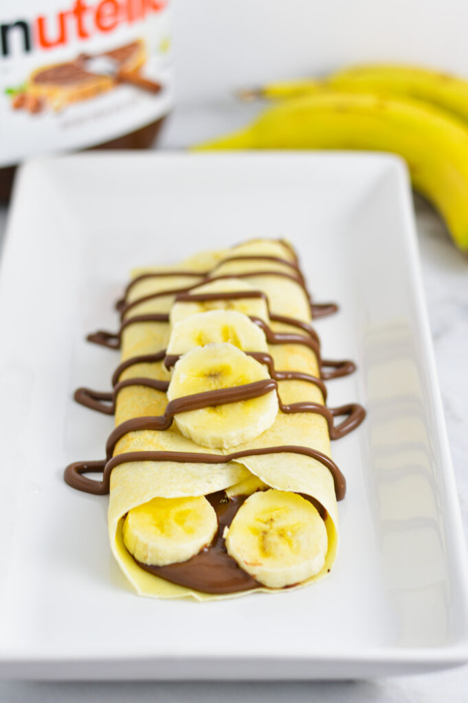 Nutella Banana Crepes on a long white plate with bananas and a jar of Nutella in the background.