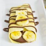 Banana and Nutella filled crepe with Nutella drizzle on a white rectangular plate.