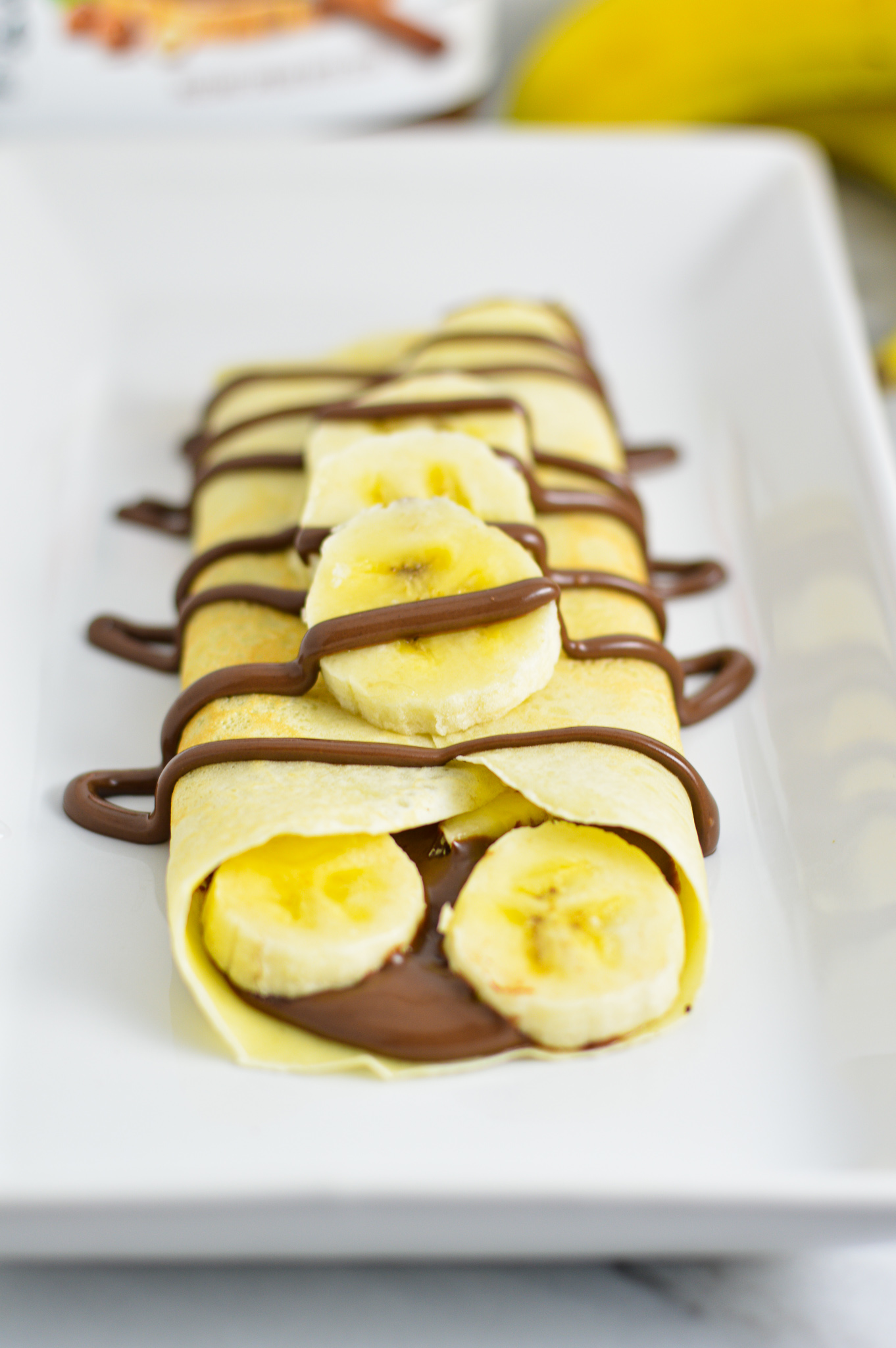 Banana and Nutella filled crepe with Nutella drizzle on a white rectangular plate.