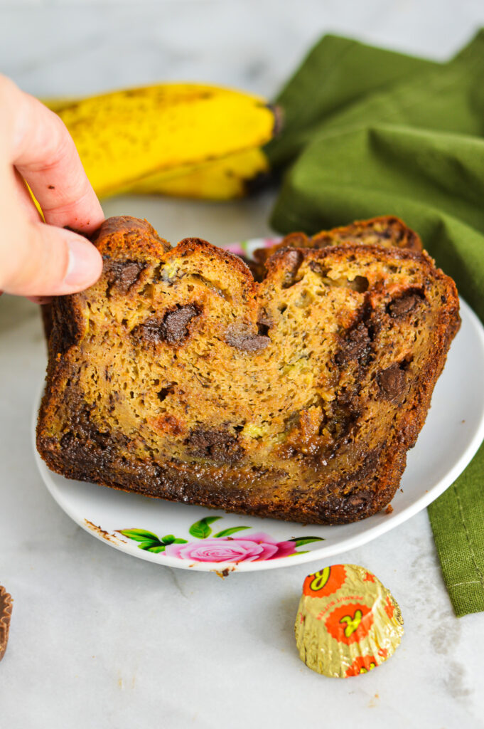 A hand grabbing a slice of Peanut Butter Cup Banana Bread with brown bananas in the background.