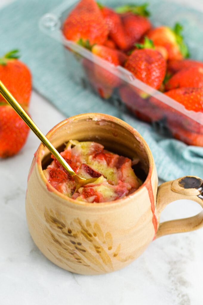 A small golden teaspoon digging into a Strawberry Mug Cake in a beige ceramic mug.