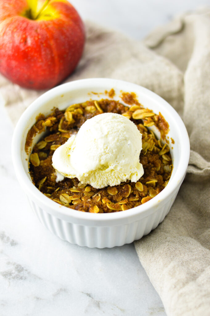 5 Minute Apple Crisp for One in a white ramekin with a red apple in the background.