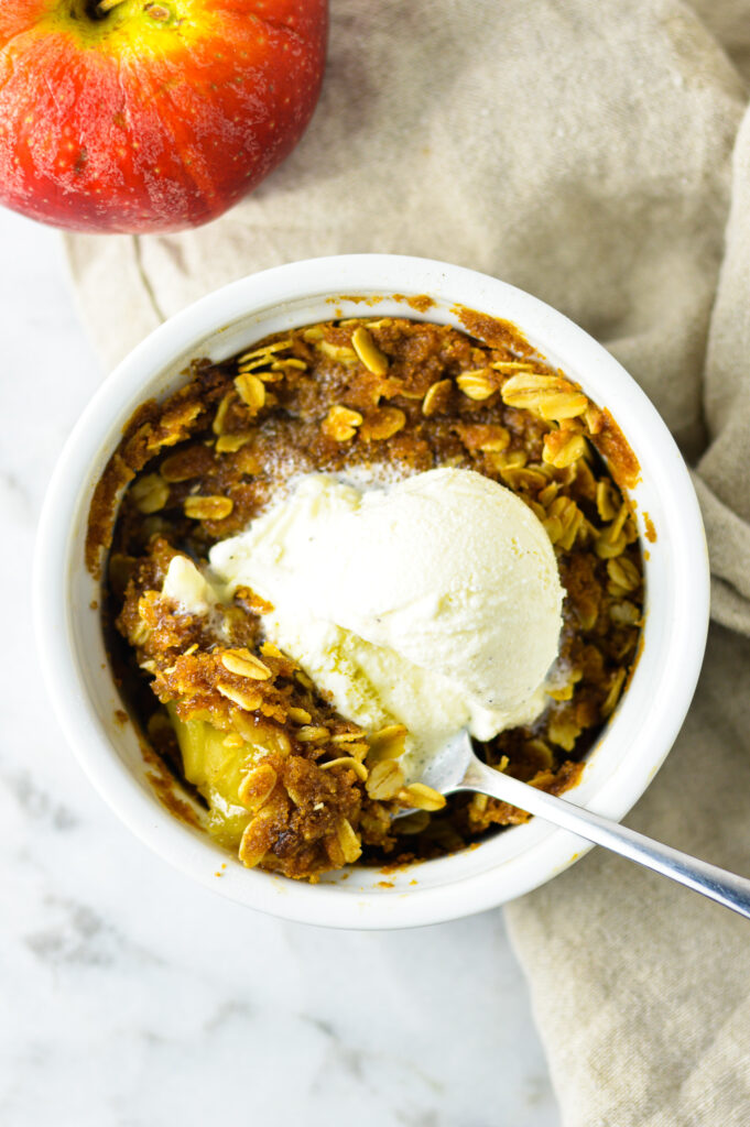 Individual Apple Crisp in a white ramekin with a scoop of vanilla ice cream and a spoon taking a spoonful.