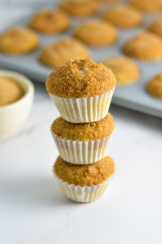 Three Churro Mini Muffins with a mini muffin pan filled with more mini muffins in the background.