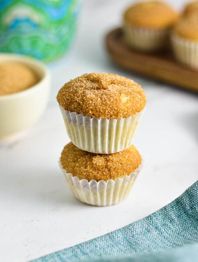 Two Churro Mini Muffins stacked with a coffee cup and a small bowl of cinnamon sugar in the background.