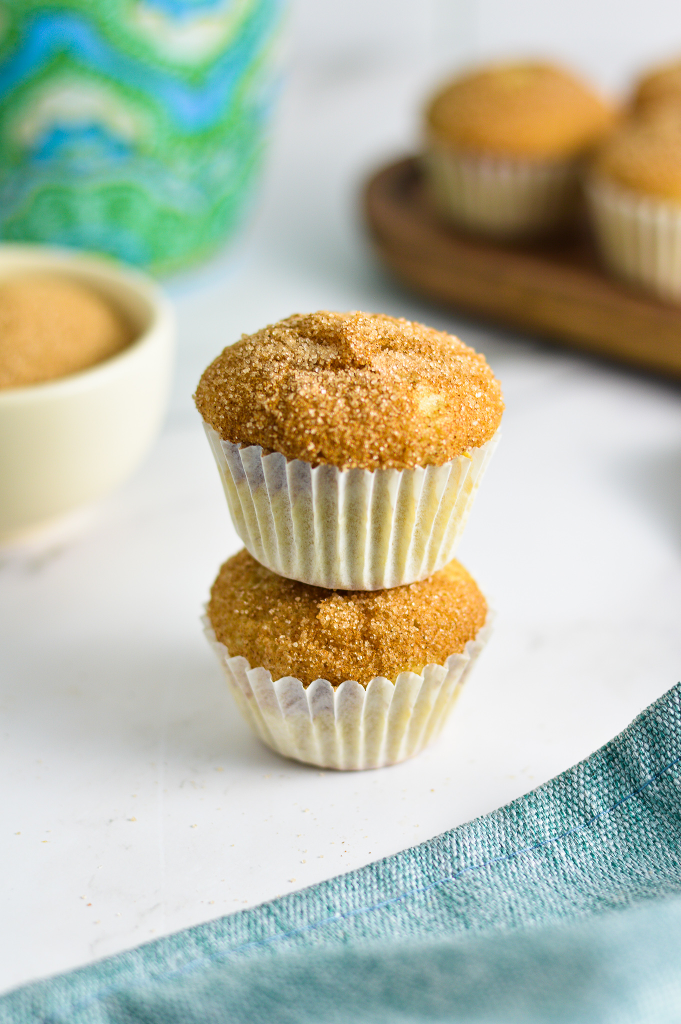 Two Churro Mini Muffins stacked with a coffee cup and a small bowl of cinnamon sugar in the background.