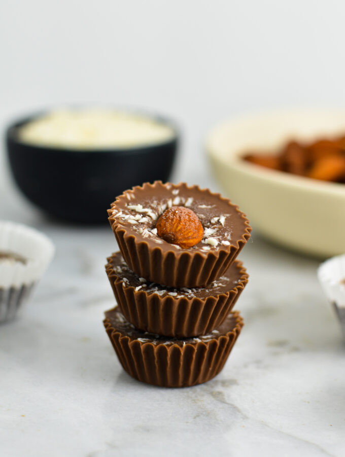 3 Almond Joy Nutella Cups stacked with a pinch bowl of coconut and a shallow white bowl of whole almonds in the background.