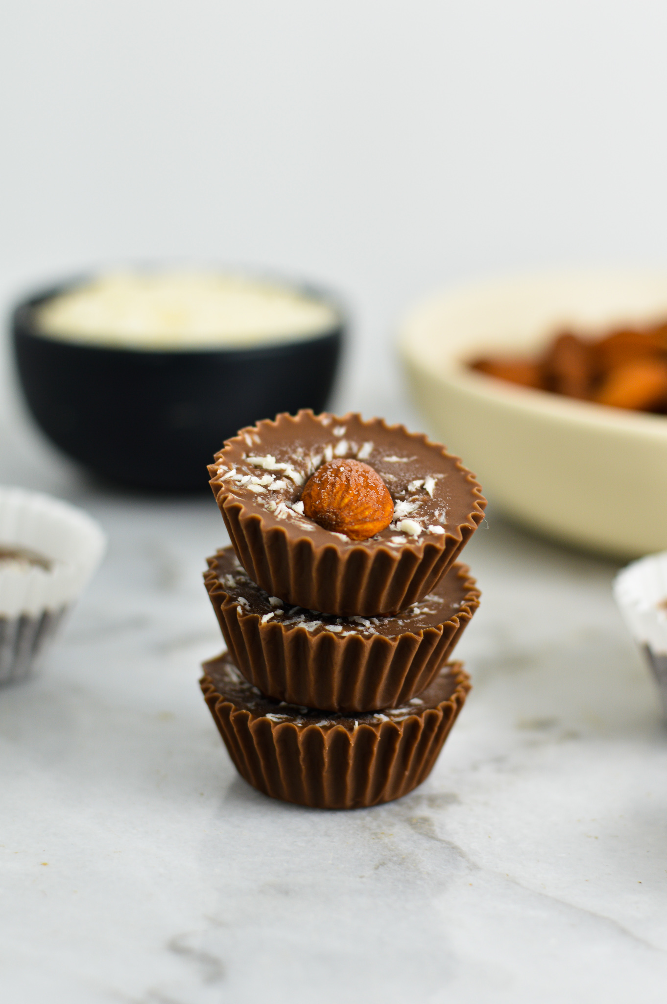 3 Almond Joy Nutella Cups stacked with a pinch bowl of coconut and a shallow white bowl of whole almonds in the background.