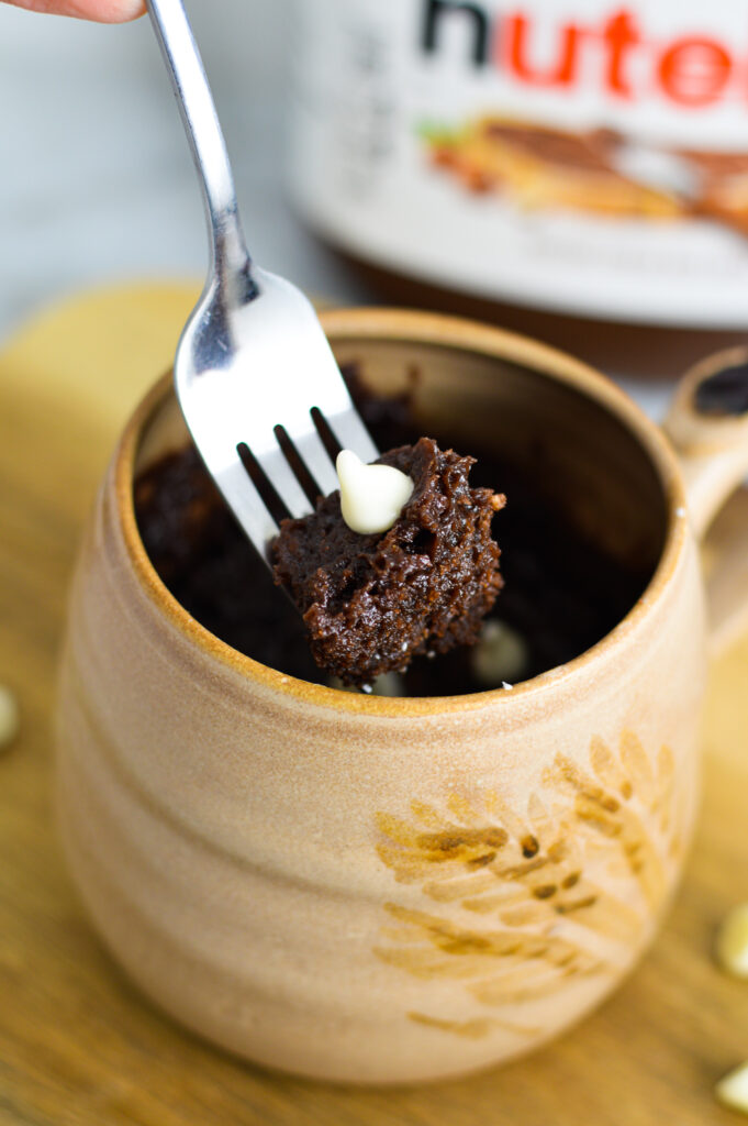 A tan ceramic mug with a fork holding a bite of White Chocolate Nutella Mug Cake.