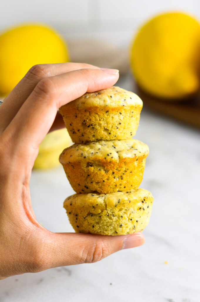 A hand holding 3 Lemon Poppy Seed Mini Muffins with medium lemons in the background.