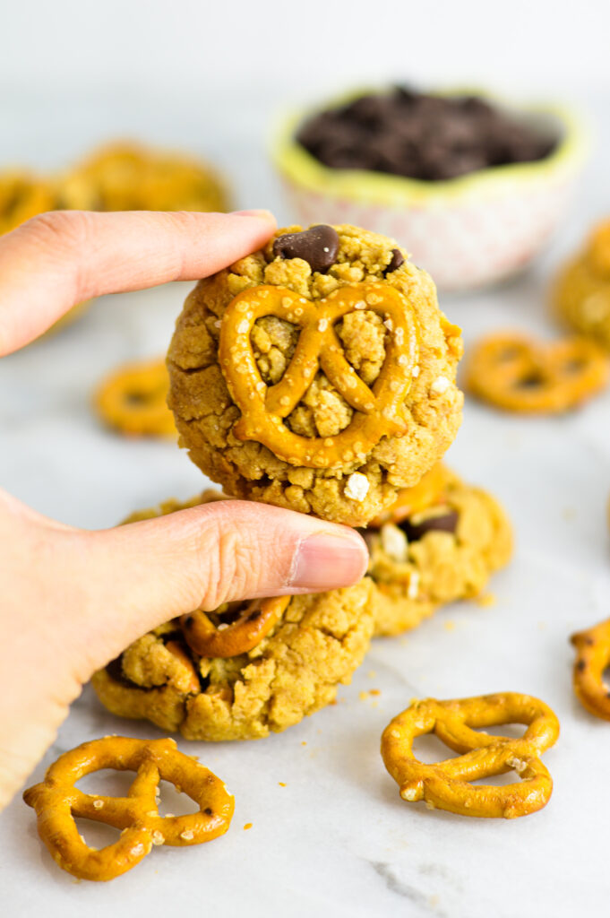 A hand holding a peanut butter chocolate chip cookie with a pretzel on top.