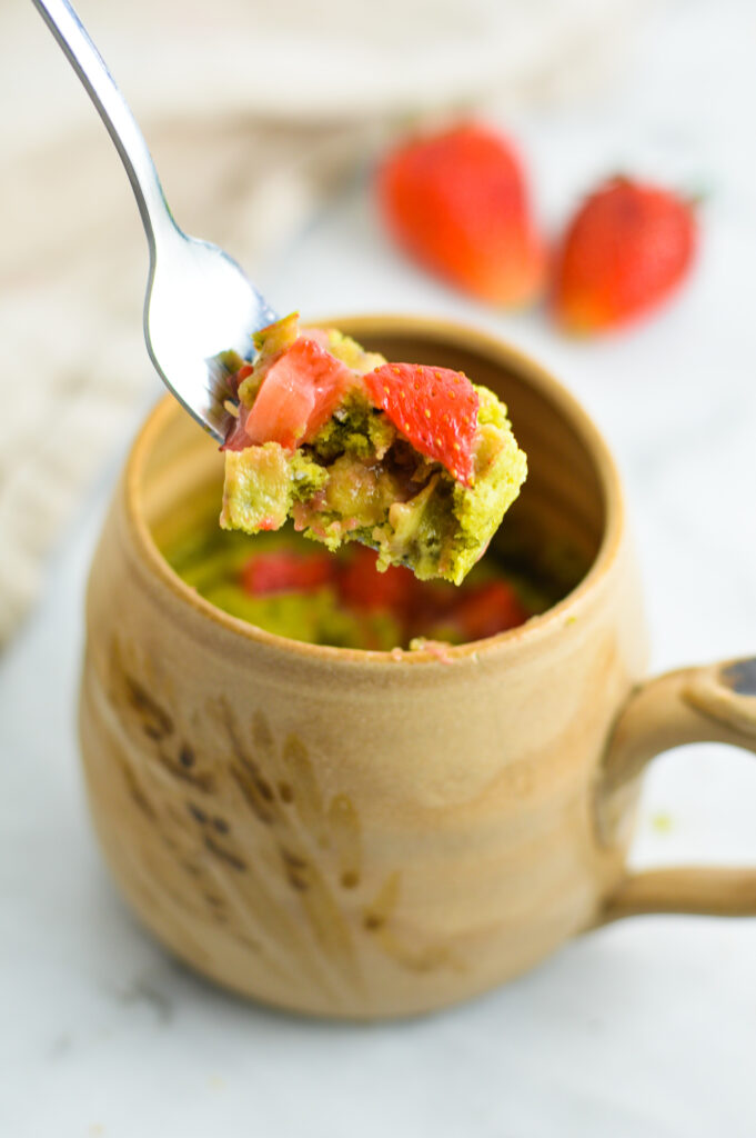 A fork full of Strawberry Matcha Mug Cake taken from a tan ceramic mug on a granite surface.