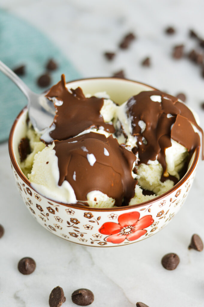 A spoon scooping ice cream topped with Vegan Magic Shell from a small bowl.