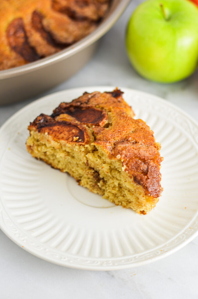 A slice of Dutch Apple Cake on a small ceramic white plate with a granny smith apple in the background.