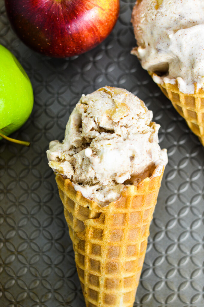 Apple Cinnamon No Churn Ice Cream in a waffle cone with granny smith and red delicious apples in the background.