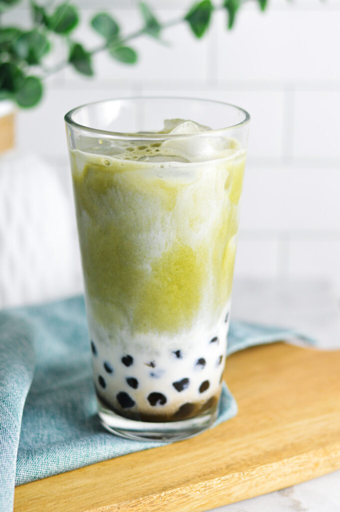 A refreshing glass of Iced Matcha Bubble Tea sits on a wooden board. The matcha and milk portions are not fully combined, with dark brown tapioca pearls peeking through the milk.