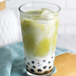 A cool glass full of ice and Iced Matcha Bubble Tea. The black tapioca pearls peek through the creamy milk, with vibrant green matcha drink swirling into the milk.