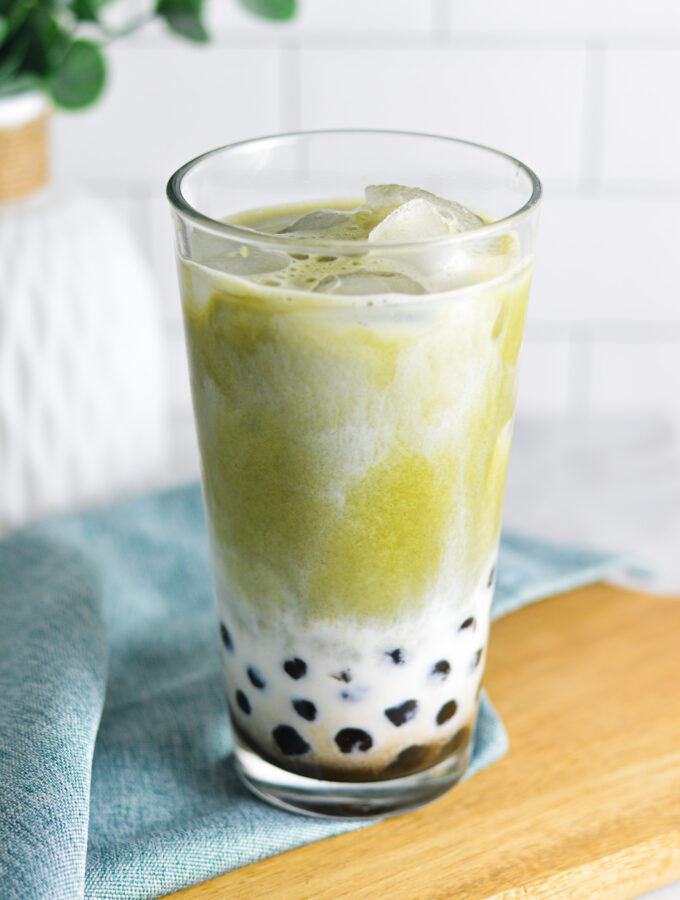 A cool glass full of ice and Iced Matcha Bubble Tea. The black tapioca pearls peek through the creamy milk, with vibrant green matcha drink swirling into the milk.