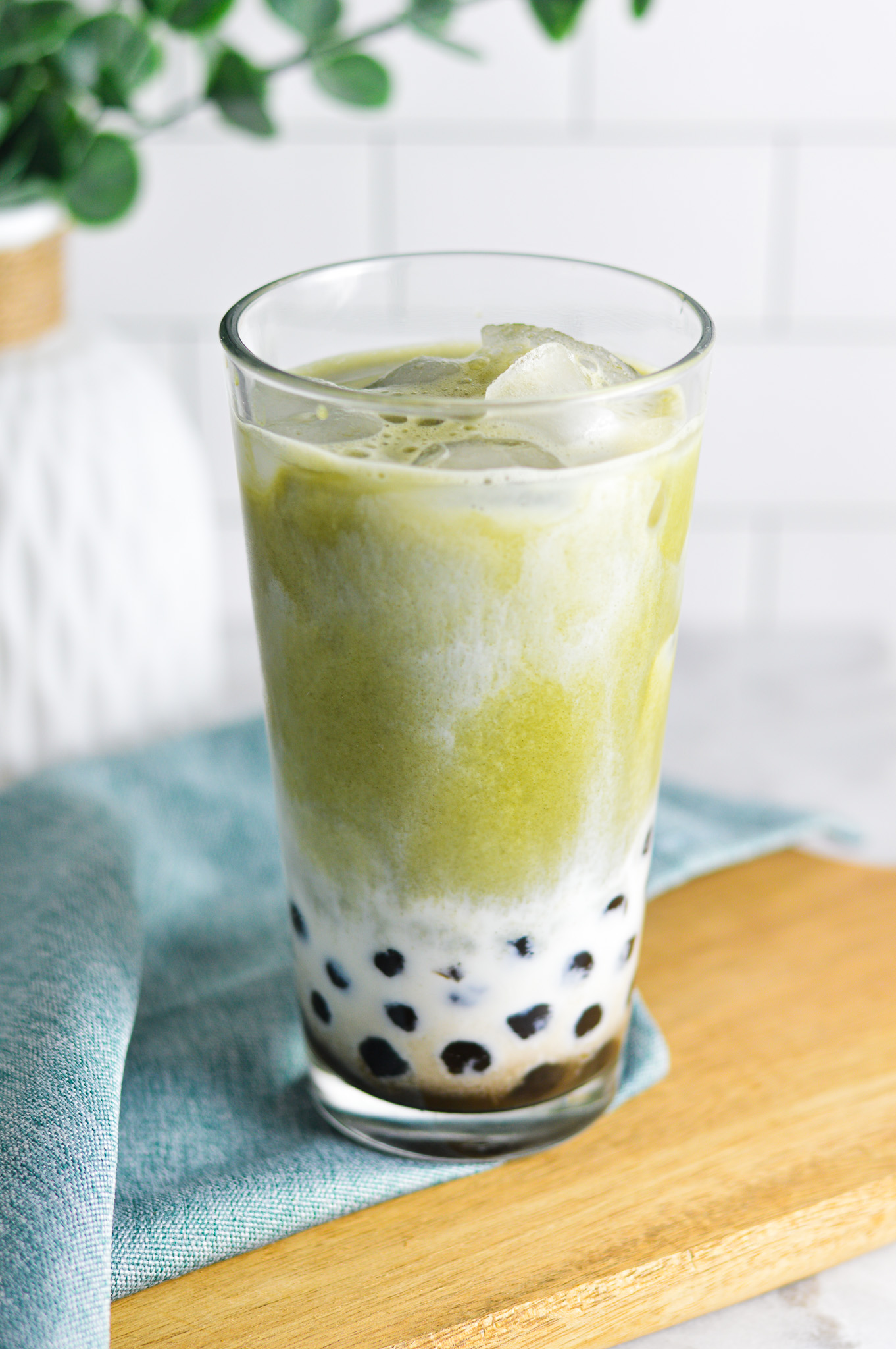 A cool glass full of ice and Iced Matcha Bubble Tea. The black tapioca pearls peek through the creamy milk, with vibrant green matcha drink swirling into the milk.