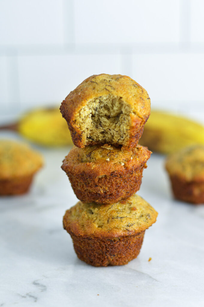 Soft Banana Mini Muffins stacked on top of each other with a ripe banana in the background.