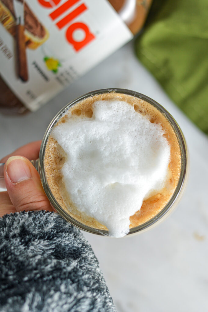 A hand holding the handle of a glass cup filled with creamy Nutella Latte.