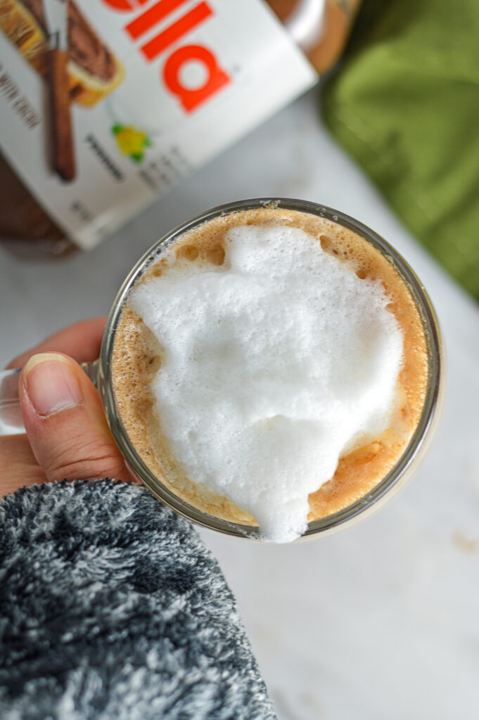 A hand holding a mug with Nutella Latte and frothed milk in it.