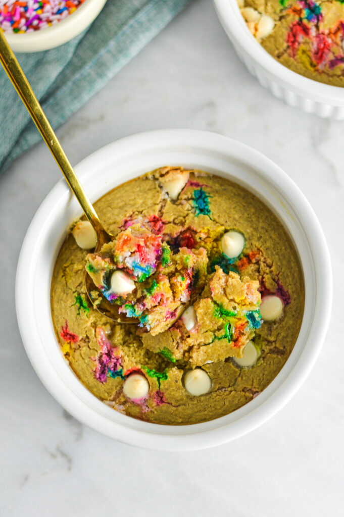 A bowl of Cake Batter Protein Blended Baked Oats with colorful sprinkles and white chocolate chips on top. A small gold spoon is taking a scoop in the center of the golden oats.