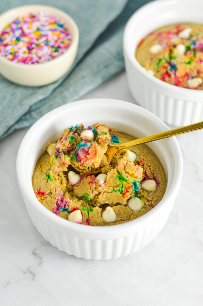 A ceramic ramekin filled with Cake Batter Protein Blended Baked Oats with a gold spoon in it showing a fluffy cakelike texture.