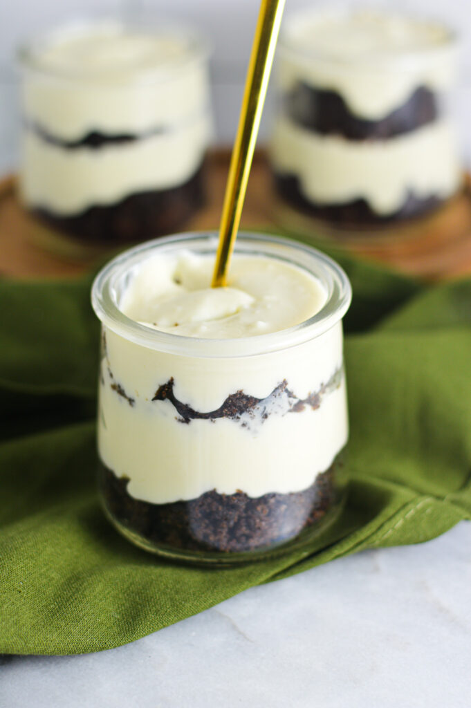 Personal sized Chocolate Cake Cheesecake Cups on a surface featuring a small wooden board and a dark green napkin. The front glass jar shows the layers of moist chocolate cake and creamy cheesecake with a gold spoon in it.