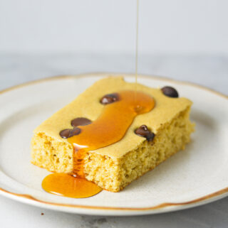 A chocolate chip filled sheet pan pancake slice on a small plate with maple syrup being drizzled on top.