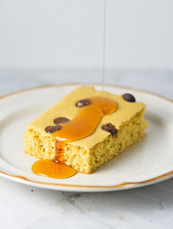 A chocolate chip filled sheet pan pancake slice on a small plate with maple syrup being drizzled on top.