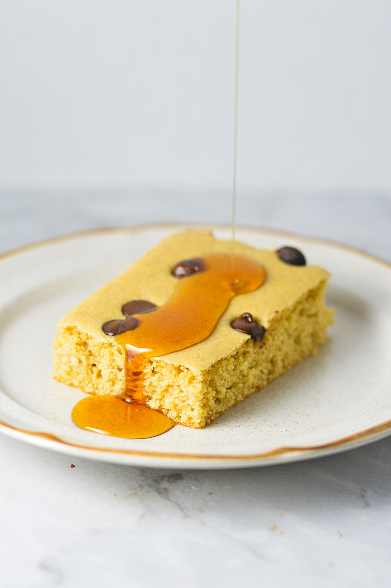 A chocolate chip filled sheet pan pancake slice on a small plate with maple syrup being drizzled on top.