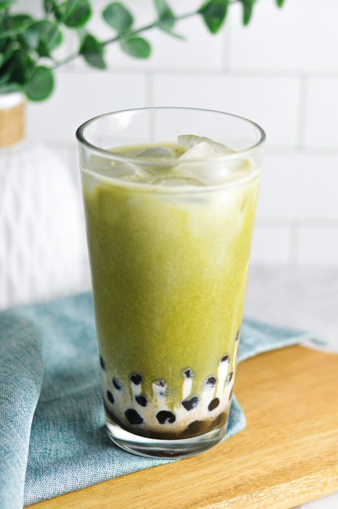 A tall clear glass filled with Iced Matcha Bubble Tea sits on a wooden surface. The glass is surrounded by a subtle sheen of condensation, suggesting that the drink is cold and refreshing.