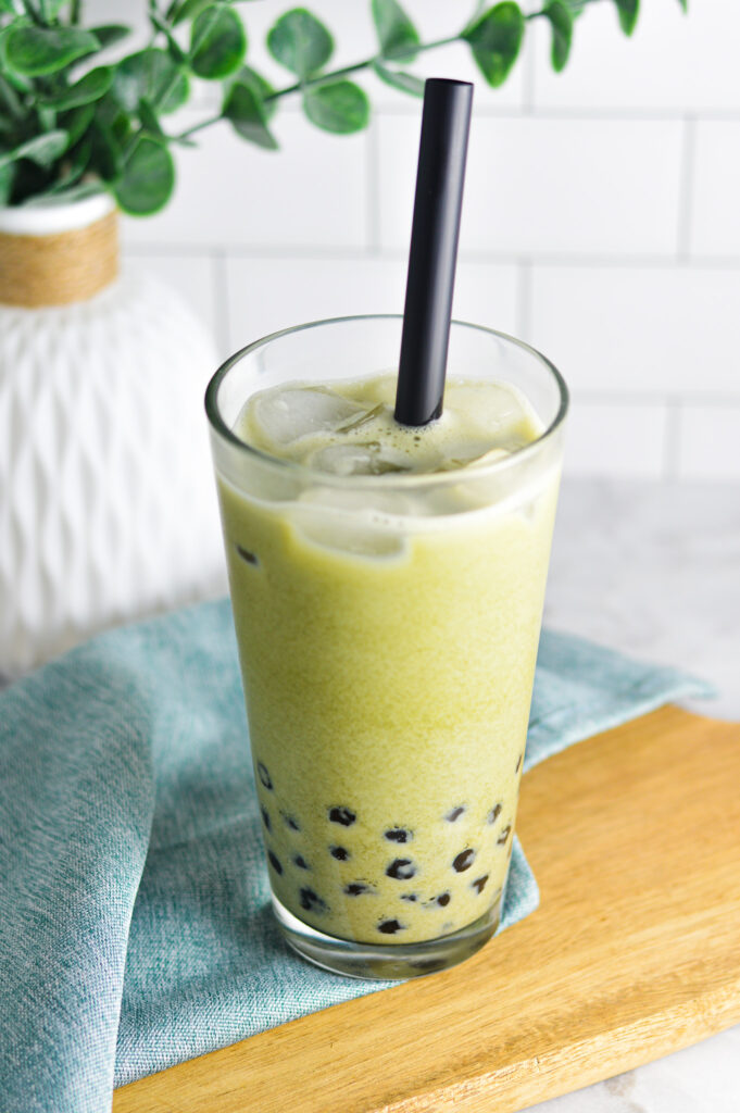 A highball glass filled with creamy Iced Matcha Bubble Tea sits on a wooden surface. A large black straw pokes through the top of the bright green drink topped with ice cubes.