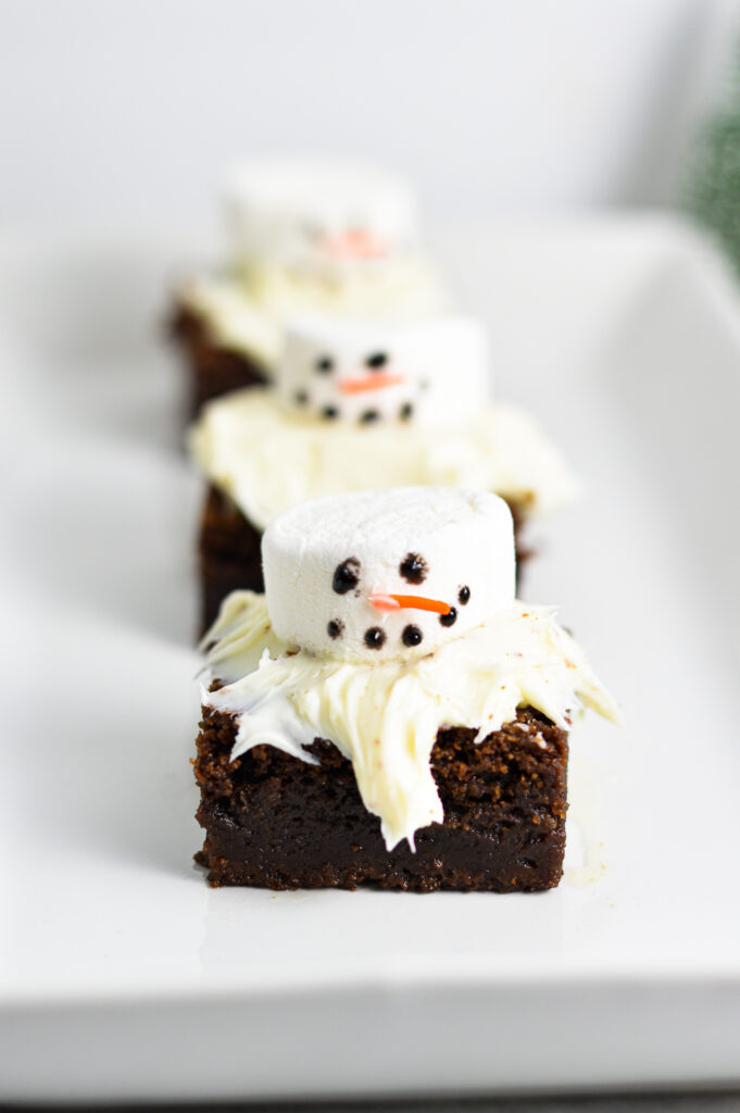 Three Melted Snowman Nutella Brownies arranged on a long white plate. An orange sprinkle is used as the snowman's nose, and black food gel made the eyes and mouth.