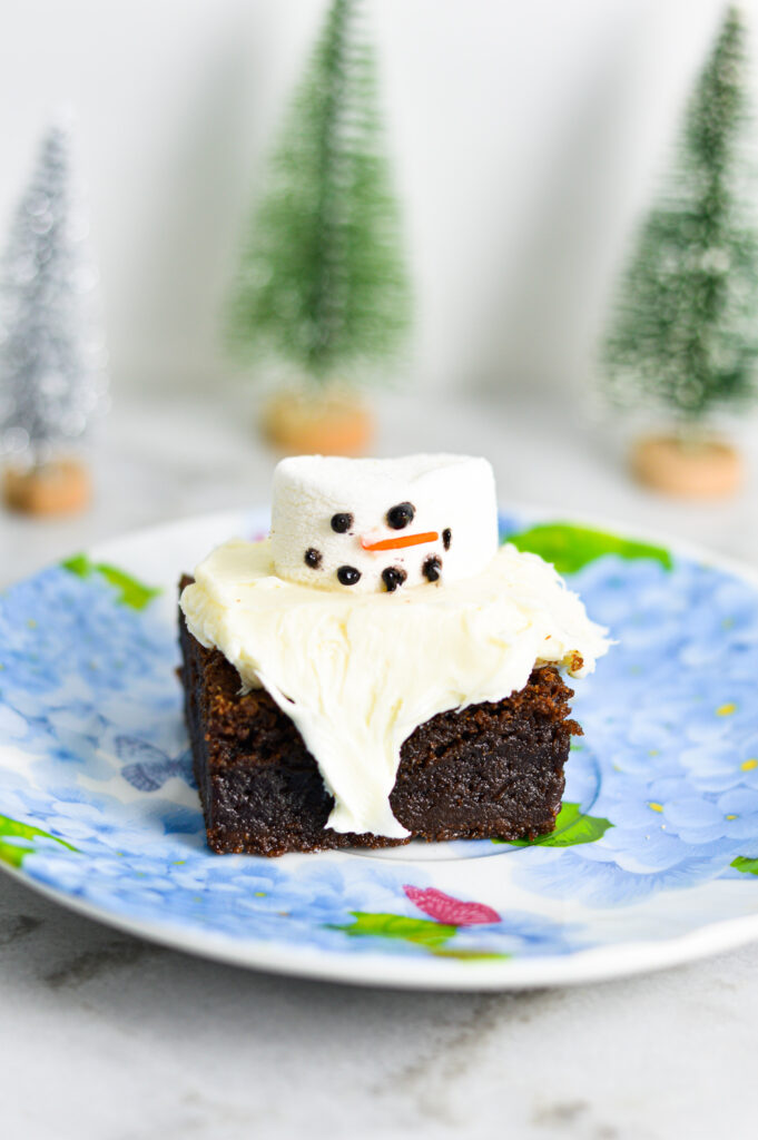 A brownie topped with a snowman decoration made from a large marshmallow, orange sprinkles, and black food gel. The brownie has a rich, fudgy texture with the cream cheese glaze spilling off the sides.