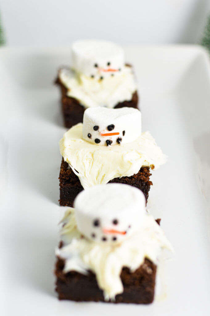 Three fudgy Nutella brownies arranged on a white plate. The fluffy marshmallows are decorated to look like snowmen with an orange sprinkle nose and black writing gel for the eyes and mouth.