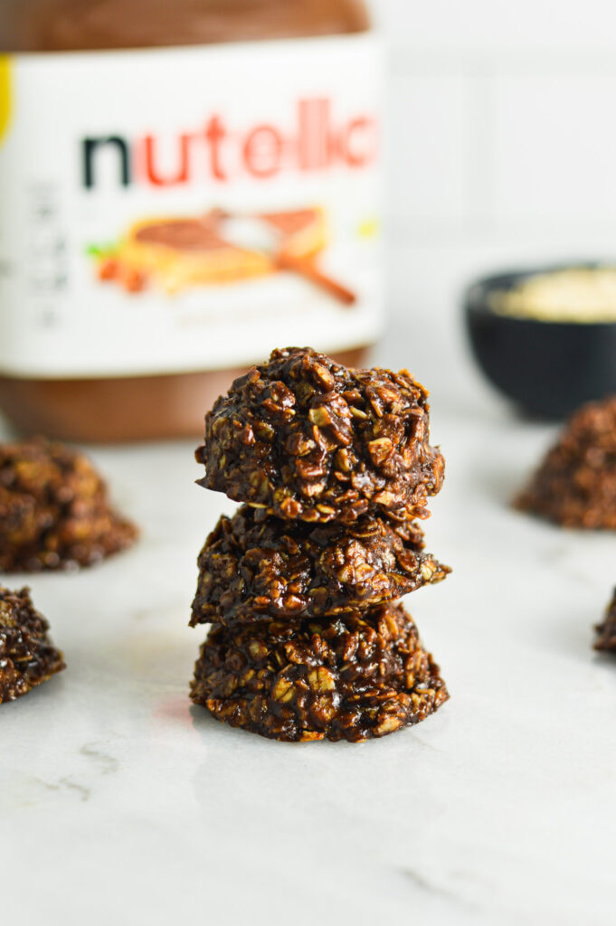 A stack of glossy No Bake Nutella Oatmeal Cookies with a jar of Nutella and a small bowl of old fashioned oats in the background.