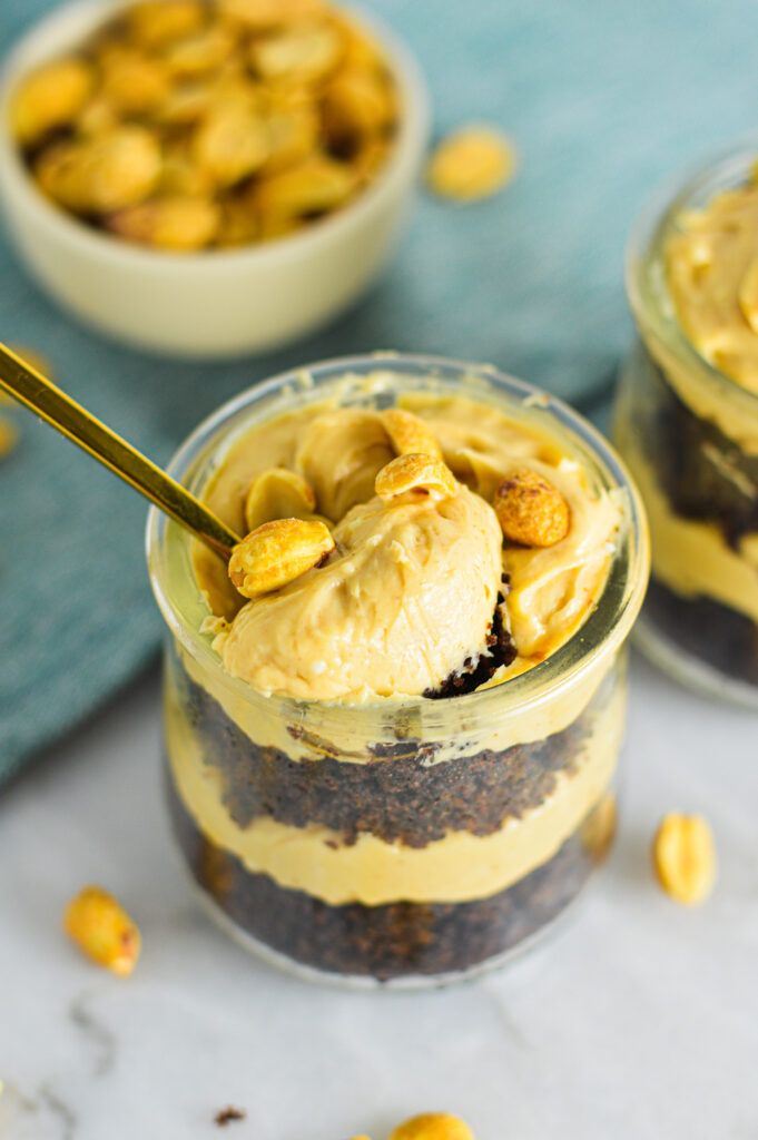 A gold spoon taking a scoop out of a Peanut Butter Chocolate Cake Cheesecake Cup. Crunchy peanuts sit atop a smooth peanut butter cheesecake with layers of rich chocolate cake. 