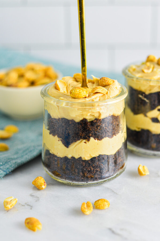 A gold spoon sticking out the top of a Peanut Butter Chocolate Cake Cheesecake Cup. There is a small bowl full of roasted peanuts in the backgroud.