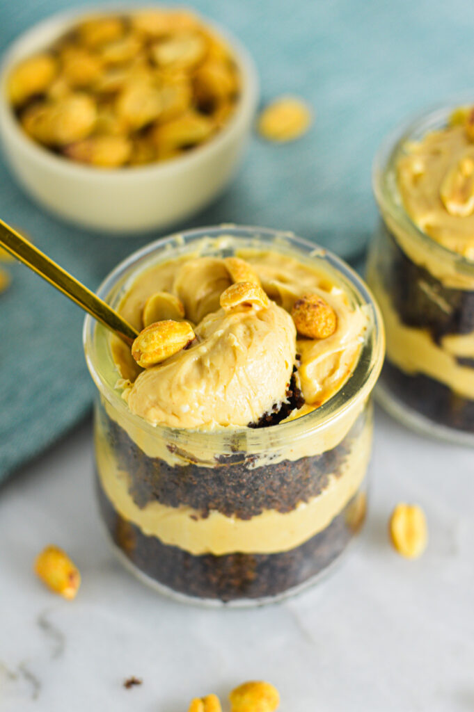 A small spoon scooping a Peanut Butter Chocolate Cake Cheesecake Cup. The layers of chocolate cake and decadent peanut butter cheesecake are showing through the small glass container.