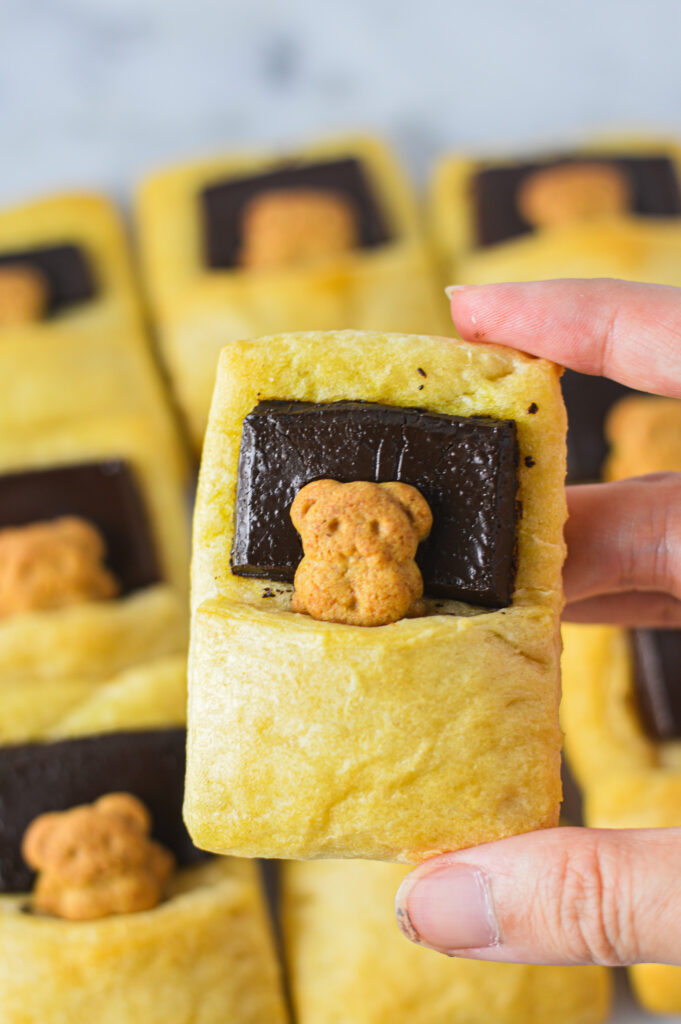 A hand holding a perfectly golden Bear in a Blanket, with more freshly baked pastries in the background.