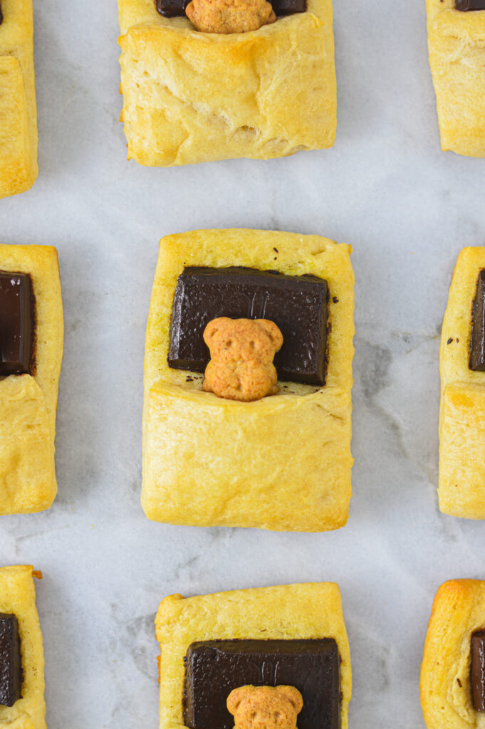 Bears in a Blanket neatly lined on a granite surface. The melted chocolate square has a graham cracker teddy bear nestled in a golden brown blanket made of baked crescent dough.