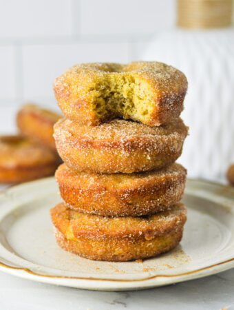 A small plate with a stack of Cinnamon Sugar Banana Bread Donuts. The top donut has a bite taken out of it revealing a fluffy cakelike texture.