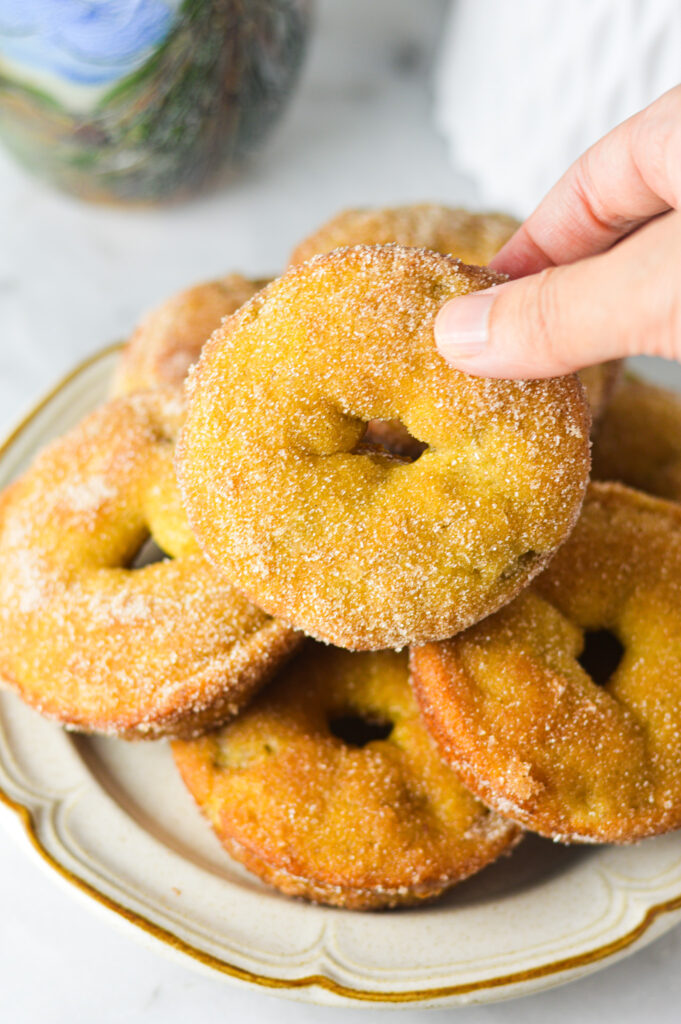 A hand taking a Cinnamon Sugar Banana Bread Donut from a plate. The light cinnamon sugar coating creates a crisp and sweet exterior.