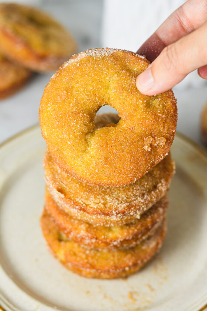 A stack of Cinnamon Sugar Banana Bread Donut with a hand taking the top donut. The texture of the donuts shows a rustic homemade feel.